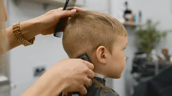 Hairdresser girl cuts the hair of blonde child with an electric trimmer machine. Process cutting hair in barber shop young girl. Men haircut and hair styling in salon. Cutting hair with trimmer. 免版税图库图片