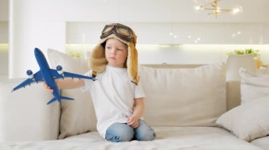 Happy little child playing with white passenger toy plane wearing goggles and pilot cap dreams of flying above clouds while sitting on sofa of apartment during day. Travel and adventure concept.