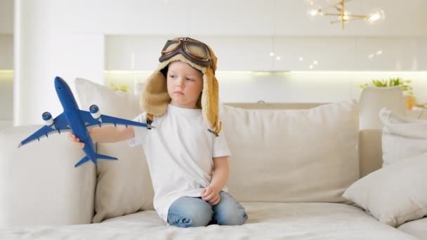 Happy little child playing with white passenger toy plane wearing goggles and pilot cap dreams of flying above clouds while sitting on sofa of apartment during day. Travel and adventure concept. — Video