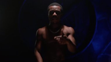 Cool African American teenager with bare, pumped-up body sings hip-hop song and emotionally gestures with his hands. Man raps in music video and sings rap song against blue underground background.