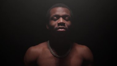 Portrait serious black man young guy looking into camera. Slow motion shot young African-American man gaze in dark studio room only one light. Man look, experience, stress in man.