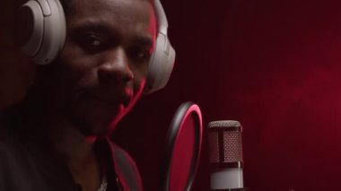 Slow motion portrait of an African American singer in recording studio with headphones on his head and studio professional microphone looks at camera and shows crossed fingers signs.