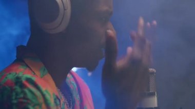 Black man portrait sings rapping into a microphone with headphones on his head, emotionally waving his arms in smoky room. Portrait of black singer in headphones recording music track in smoky room.
