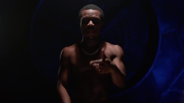 Cool African American teenager with bare, pumped-up body sings hip-hop song and emotionally gestures with his hands. Man raps in music video and sings rap song against blue underground background. — Stockvideo