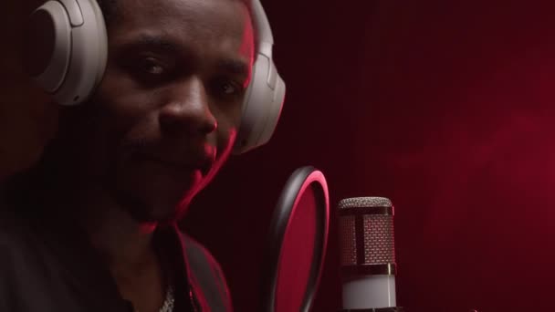 Slow motion portrait of an African American singer in recording studio with headphones on his head and studio professional microphone looks at camera and shows crossed fingers signs. — Video
