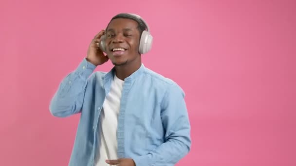 Slow motion portrait black man teenager 20 years, in denim shirt with headphones on his head listens to his favorite music, sings words of song, waves his hands to rhythm of music on pink studio. — Αρχείο Βίντεο