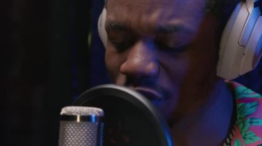 Portrait young handsome African American singer emotionally working in studio microphone with headphones on his head in recording studio against background of sound absorbing material.