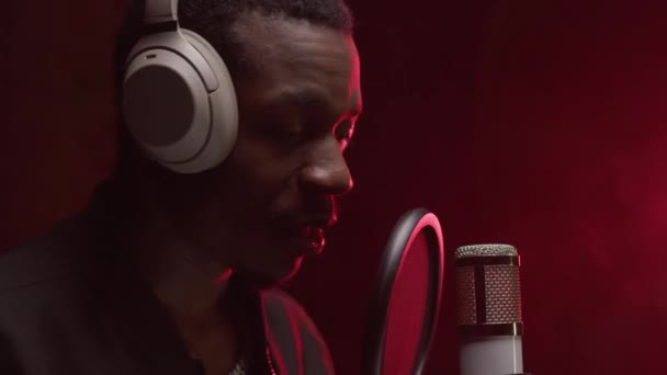 African American singer performs song while standing in recording studio with headphones on his head and sings into microphone, emotionally waving his arms. Rap singer singer sings in microphone. — Video