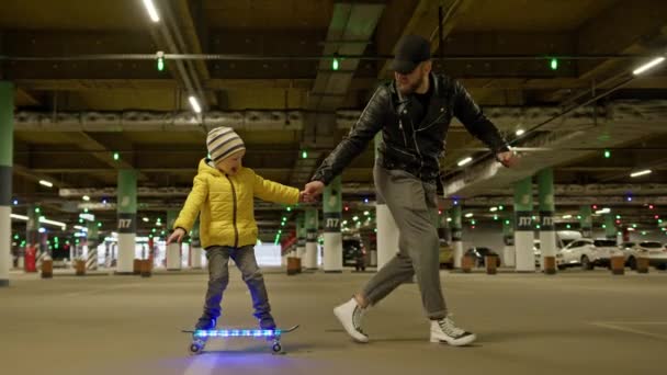 Happy boy in yellow jacket with his father learns to ride glowing skateboard in an underground parking lot. Father teaches his son to skateboard in evening in an underground parking lot. Happy family. — Vídeos de Stock