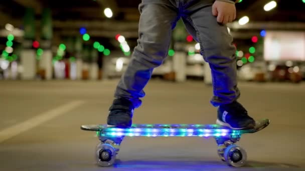 Close-up of child legs riding on skateboard glowing led with lights in an urban background. Boy rides glowing skateboard in evening in an underground parking lot. Child learns to ride skateboard. — Videoclip de stoc
