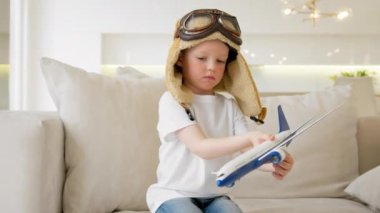 Cheerful portrait child plays with modern airplane sitting on couch of house. Boy in pilot hat controls toy plane flying in different directions. Child dreams of flying an airplane, traveling by air.