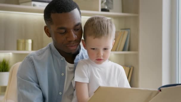Black skin father teaches his 4-year-old adopted caucasian son to read. Young African-American father reads book to his adopted child of European appearance sitting at home on chair in bright room. — Vídeo de Stock