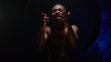 Handsome black African American man with pumped naked torso sings song emotionally waving his arms on blue underground background. Shooting clip of rap artist. Man sings rapping song.