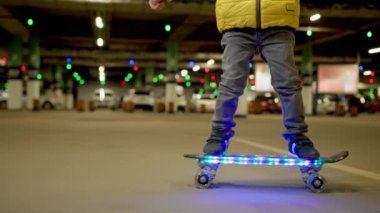 Boy rides glowing skateboard in evening in an underground parking lot. Child learns to ride skateboard. Close-up of child legs riding on skateboard glowing led with lights in an urban background.