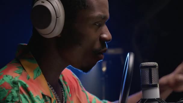 Man sings rap into microphone with headphones on his head emotionally waving his hands on blue light. Portrait black person singer in headphones recording music track in professional recording studio. — 图库视频影像