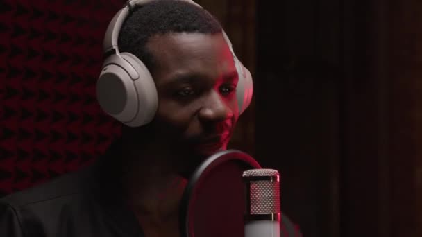 Black man singer in headphones recording music track in professional recording studio. Man sings rap song into microphone with headphones on his head against background noise-absorbing foam rubber. — Video