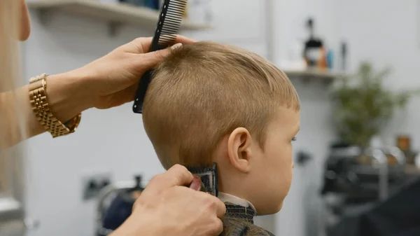 Hairdresser girl cuts hair of blond child with hair clipper in barber shop. Mens styling and hair cutting in salon. Cutting hair with trimmer. Barber gets haircut in barbershop. Stockfoto