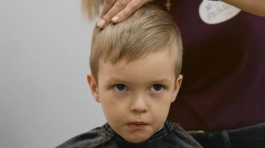Hairdresser girl cuts the hair of blonde child with an electric trimmer machine. Process cutting hair in barber shop young girl. Men haircut and hair styling in salon. Cutting hair with trimmer.