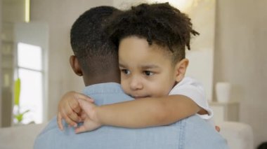 Happy childhood of 3-year-old multiracial boy in arms of black father, happy childhood of mixed-race boy. Child looks into camera hugging his father sitting in his arms at home.