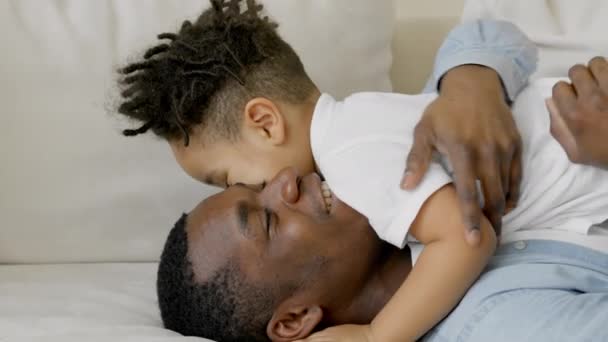 Multiracial Boy and his father ride ropes and play together. Concept of family fun pastime. Happy joyful African American dad hugs and tickles laughing multi-race little son on couch in living room. — Vídeo de Stock