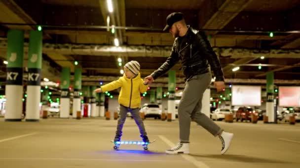 Father supports the child who learns to ride skateboard in evening at parking lot. Happy father and son family, father teaches young son to ride glowing led skateboard on an underground parking lot. — ストック動画