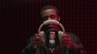 Black man singer in recording studio stands near studio microphone and puts gray headphones on his head against soundproof wall. Afro American is preparing to record song.