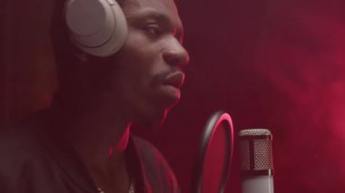 African American Male Singer Recording New Track, Slow Motion. Music Production Concept. Young Black Guy In Professional Headphones Singing Song To Microphone At Sound Record Studio With Red Lighting.