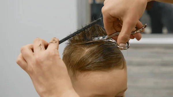 Process of hairdresser cutting hair. Blond caucasian boy is sitting in barber shop and hairdresser girl is cutting his hair. Child gets fashionable haircut, hairdresser cuts child hair with scissors.