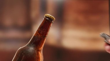 Close-up man hand opens lid fresh light beer in sweating from cold dark brown glass bottle with lid opener on blurry background. Slow-motion video light lager is opened with help of bottle opener.