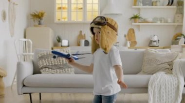 Happy child in aviapilot hat is holding toy airplane in his hands, circling with an airplane and dreaming of flying in clouds. Child aspires to fly high in clouds playing with an airplane at home.