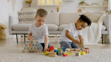Two small children of different ethnic groups play together with wooden cubes, build tower and put words out of cubes sitting on floor in bright room. Friendship game together small children at home.