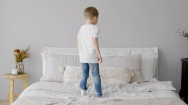 Happy blonde 4-year-old child is having fun jumping on bed, rejoicing and enjoying childhood and freedom at bedroom. Carefree childhood of small child jumping up on mattress of bed in bright room.