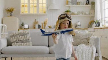 Happy child in aviapilot hat is holding toy airplane in his hands, circling with an airplane and dreaming of flying in clouds. Child aspires to fly high in clouds playing with an airplane at home.
