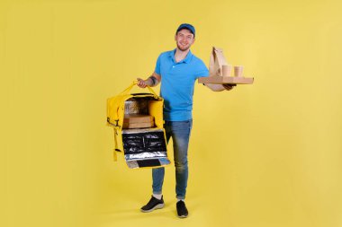 Smiling young handsome Caucasian guy delivering food orders to home and office in blue uniform holds an order for pizza and drinks in cardboard packages and thermos bag on his shoulder on yellow back.