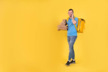 Delivery guy in blue uniform is walking talking on phone holding an order with food from restaurant and talking to customer behind his back he has delivery guys refrigerator bag on yellow background.