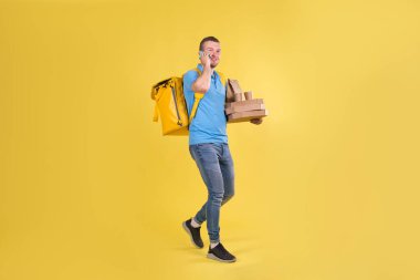 Delivery guy in blue uniform is walking talking on phone holding an order with food from restaurant and talking to customer behind his back he has delivery guys refrigerator bag on yellow background.