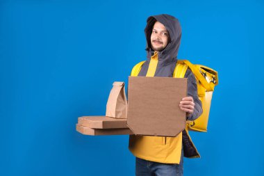Koyu renk saçlı erkek kurye restoranlardan evinize ya da işyerinize sırtında torbayla yemek getiriyor. Kurye mavi arka planda yiyecek pizzası içeren bir ekolojik paket tutuyor.