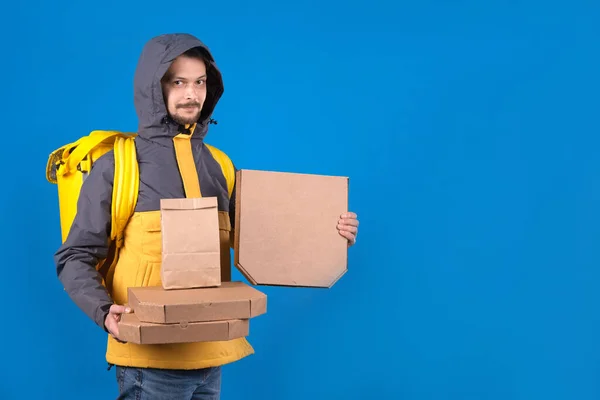 Dark-haired male courier delivering food from restaurants to your home or office in yellow clothes with bag on your back. Courier is holding an ecological package with food pizza on blue background