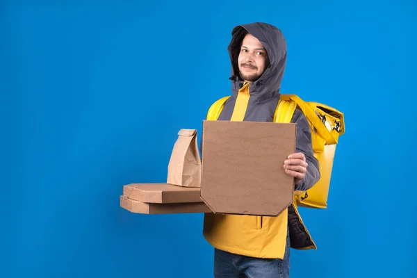 Dark-haired male courier delivering food from restaurants to your home or office in yellow clothes with bag on your back. Courier is holding an ecological package with food pizza on blue background