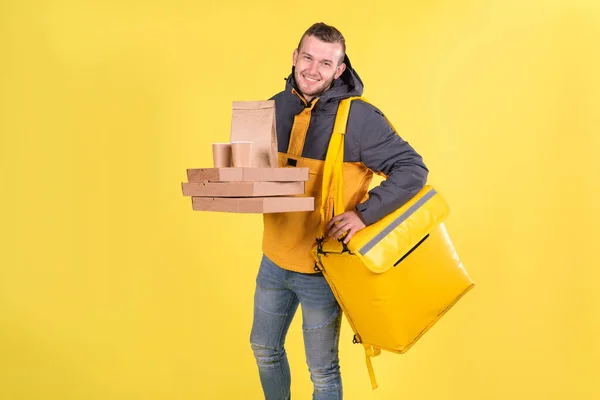 Cute caucasian food delivery courier smiles and looks ahead, holding food order in his hands and yellow refrigerator bag on yellow background hangs on his shoulder. Food delivery to home or office
