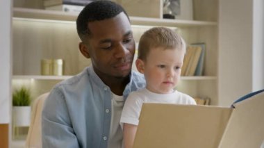 Young African-American father reads book to his adopted child of European appearance sitting at home on chair in bright room. Black skin father teaches his 4-year-old adopted caucasian son to read