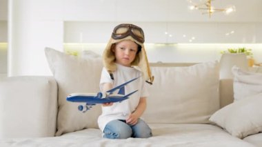 Child dreams of flying an airplane, traveling by air. Cheerful child plays with modern airplane sitting on couch of house. Boy in pilot hat controls toy plane flying in different directions