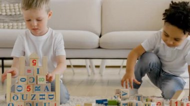 Friendship game together small children at home. Two small children of different ethnic groups play together with wooden cubes, build tower and put words out of cubes sitting on floor in bright room