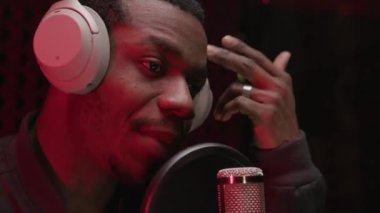 Close up portrait young African American handsome stylish guy standing at microphone in sound studio and singing on red lights