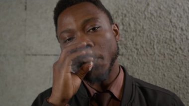 Nicotine addiction in black skin man standing near wall and smoking cigarette exhaling smoke. Portrait African-American man puts cigarette to his mouth and inhales smoke into his lungs and exhales