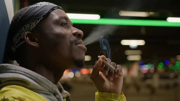 African american man smokes cigarette inhaling harmful smoke with nicotine while standing near wall in an underpass. Portrait smoking black skin teenager smoking marijuana and exhaling smoke