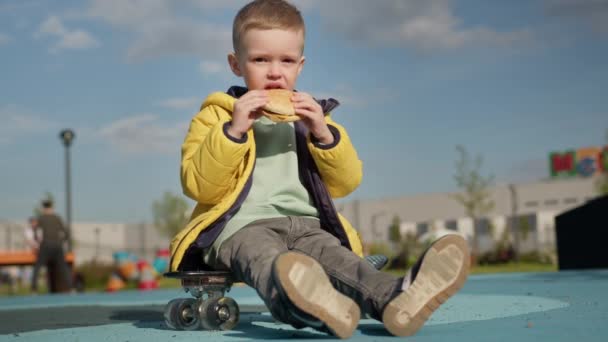 Blond Haired Child Yellow Jacket Sits Skateboard Eats Beef Burger — стоковое видео