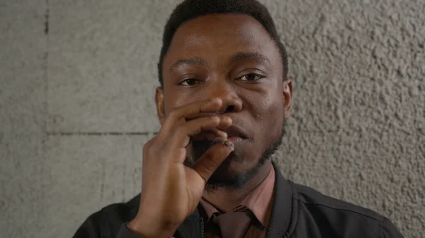 Nicotine addiction in black skin man standing near wall and smoking cigarette exhaling smoke. Portrait African-American man puts cigarette to his mouth and inhales smoke into his lungs and exhales