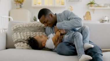 Affectionate african family young adult father tickling little adorable child son having fun laughing relaxing on sofa, afro american dad playing with small kid boy bonding cuddling together at home