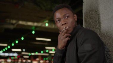 Black skin guy smokes a marijuana cigarette in an underpass against wall. Nicotine addiction, medicinal marijuana. Portrait African-American man in shirt and tie smoking standing against gray wall